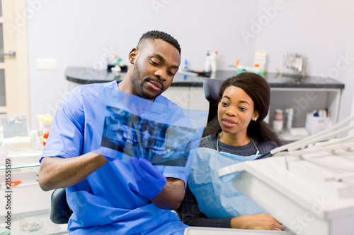 A young African man dentist in a blue medical suit and blue gloves shows the patient a Panoramic Dental X-ray of the jaws
