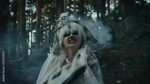 Terrifying female faun is waving by huge axe in forest. She is looking at camera in darkness, aggressive and scary