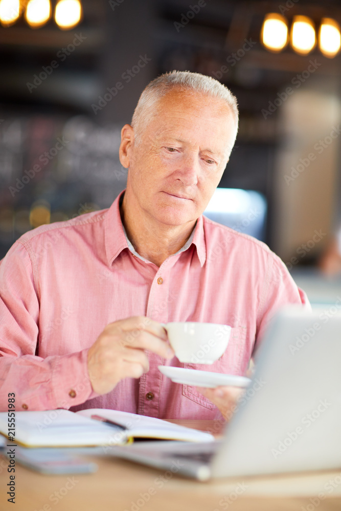 Serious aged businessman with cup of tea or coffee siting in cafe in front of laptop Serious aged businessman with cup of tea or coffee siting in cafe in front of laptop