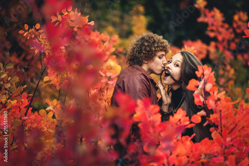 cheerful couple is naughty and shows emotions between red trees of autumn. curly-haired mustachioed man kisses, and girl makes mustache out of her hair.