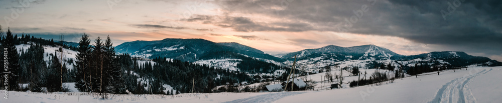 Snow-capped houses in mountains Carpathians Ukraine. On background Christmas tree in forest. Winter nature. Landscape. Top side view. Long edge. Panorama. Bukovel. Beautiful sunset over a wide valley.