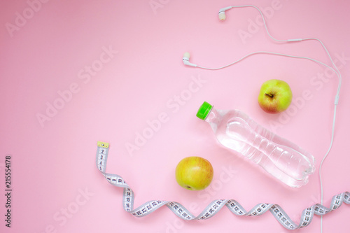Fitness flat lay. Healthy lifestyle and sport concept. Tape measure, white earphones, apples and bottle of water on a pink background.