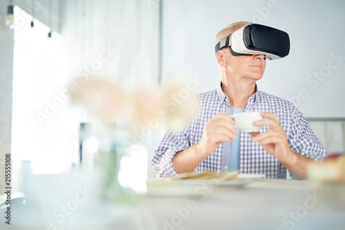 Aged restired man with cup of tea sitting by table and watching virtual webcast in vr goggles © pressmaster