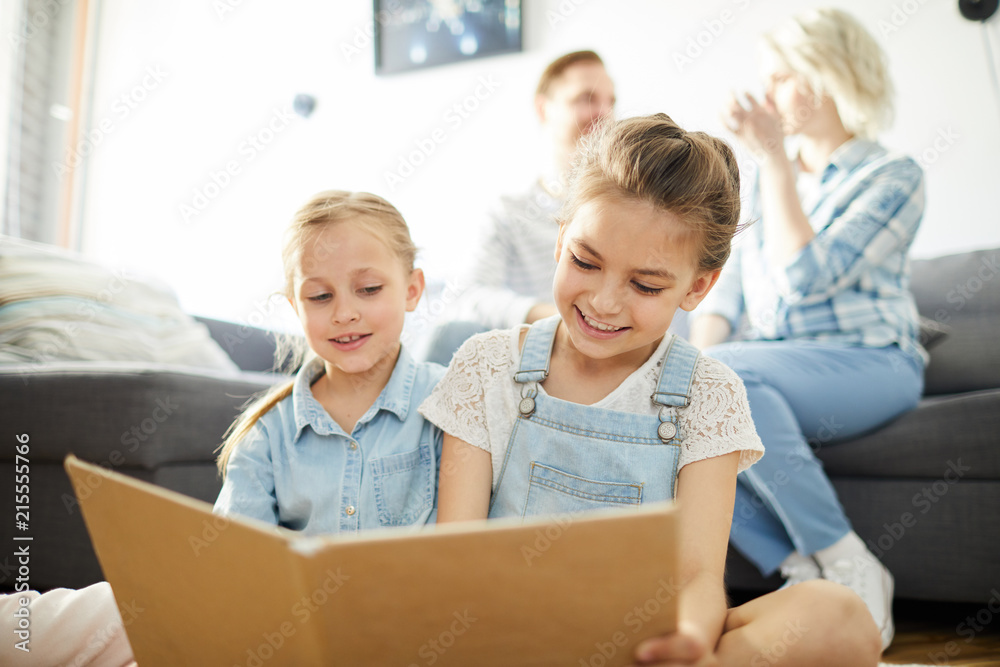 Two happy little girls reading book while sitting on the floor on background of their parents Two happy little girls reading book while sitting on the floor on background of their parents