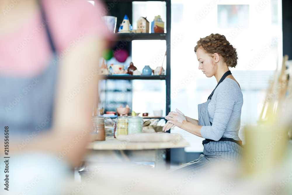 Young woman sitting by table in workshop and preparing workpieces for earthenware items Young woman sitting by table in workshop and preparing workpieces for earthenware items