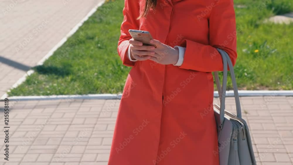 Unrecognizable woman in red coat waits for someone and checks her phone, texting. Close-up hands. Unrecognizable woman in red coat waits for someone and checks her phone, texting. Close-up hands.