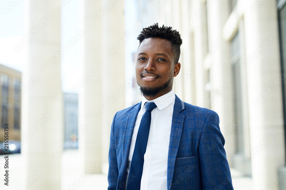 Happy young African-american employee in suit and tie looking at you in urban environment by modern business center