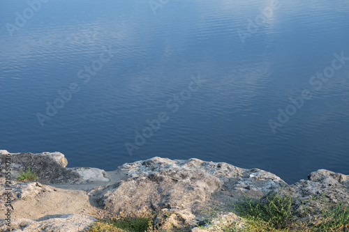 Calm Water View from the Rocky Shore Reflecting Clouds and Blue Sky