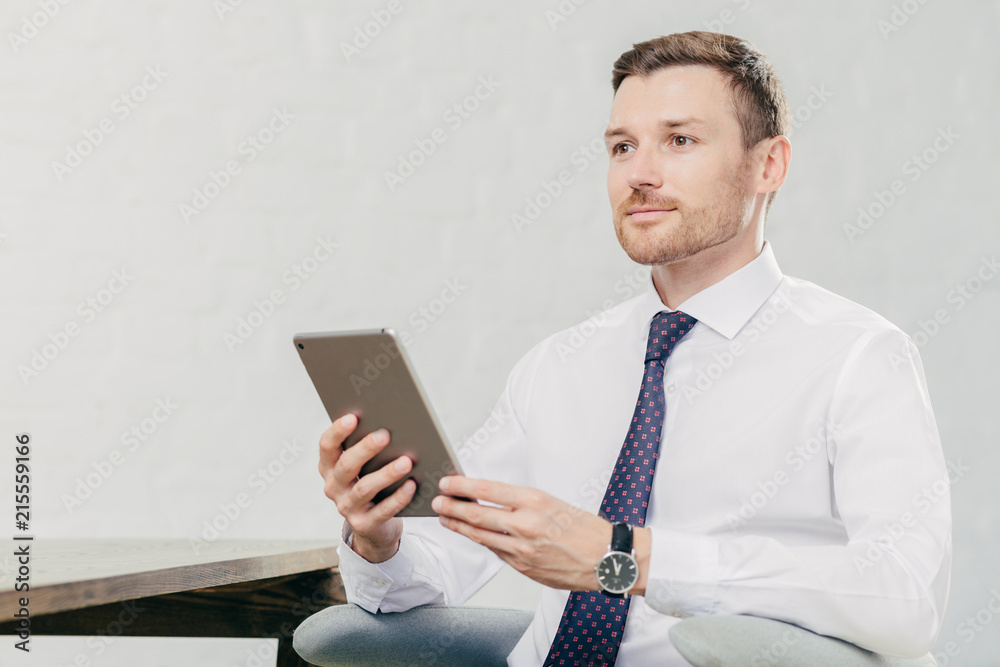 Thoughtful Caucasian male laywer thinks about something, holds modern tablet computer, wears white shirt with black tie, sits at his cabinet, waits for client. People, work, technology concept Thoughtful Caucasian male laywer thinks about something, holds modern tablet computer, wears white shirt with black tie, sits at his cabinet, waits for client. People, work, technology concept