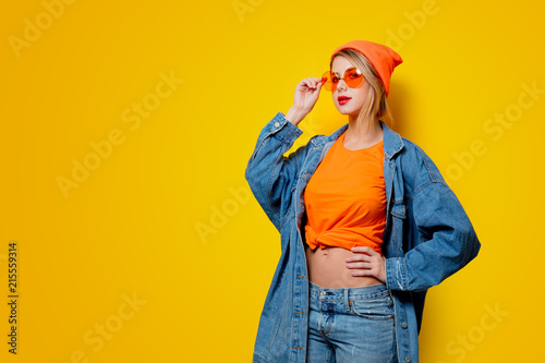 Young style girl in jeans clothes with orange glasses on yellow background. Clothes in 1980s style