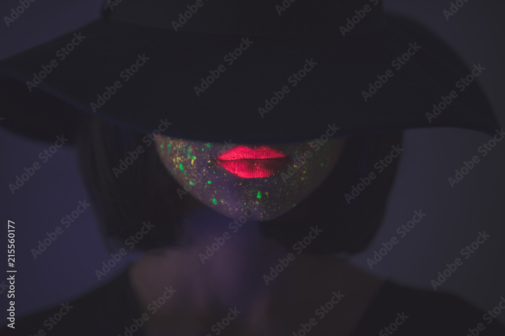 Young girl in hat with fluorescent paint on lips and face. Studio shot.