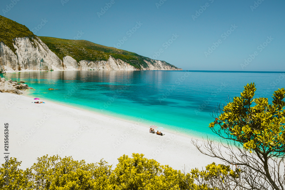 Amazing Fteri beach lagoon, Cephalonia Kefalonia, Greece. Tourists under umbrella relax near clear blue emerald turquise sea water. White rocks in background