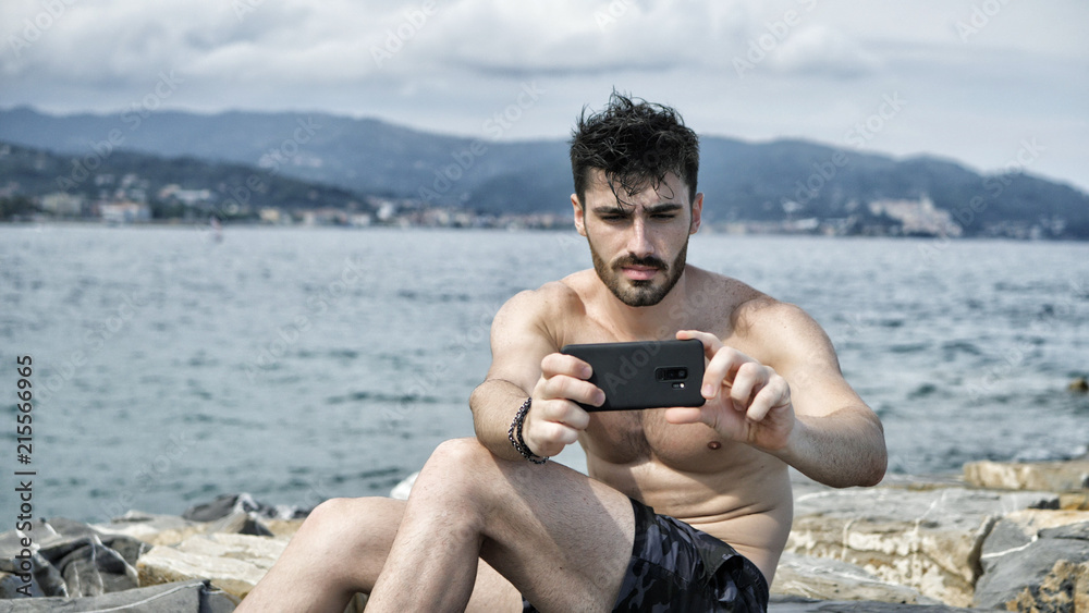 Young handsome man at the seaside using smartphone to take shots and film footage of the sea or ocean Young handsome man at the seaside using smartphone to take shots and film footage of the sea or ocean