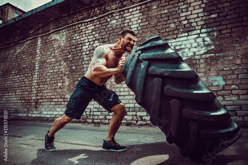 Obraz na plátne Muscular bearded tattooed fitness shirtless man moving large tire in street gym