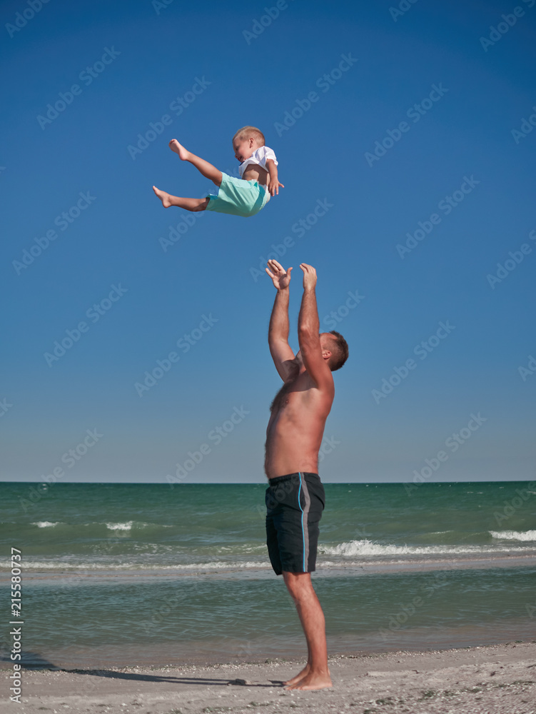 Obraz premium Dad and Baby playing on seaside in summertime. Happy family and happy childhood concept. Little boy having fun with Daddy on beach. Dad throwing and catching up a kid into air. Summer rest concept