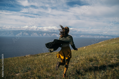 Russian girl in a coat is running across the field, there is happiness. the girl is having fun. Rear view of girl running on the background of blue sky.