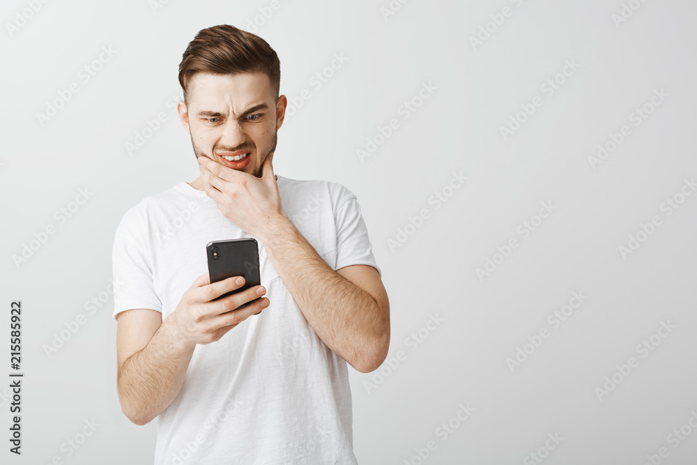 Guy send message to wrong person feeling anxious and concerned rubbing beard frowning and grimacing with opened mouth staring shook, worried at smartphone screen cannot delete mistake over gray wall Guy send message to wrong person feeling anxious and concerned rubbing beard frowning and grimacing with opened mouth staring shook, worried at smartphone screen cannot delete mistake over gray wall