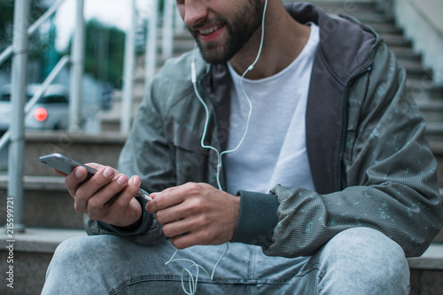 Song and the city. Close-up part of young man in headphones using his smartphone while sitting against industrial city view