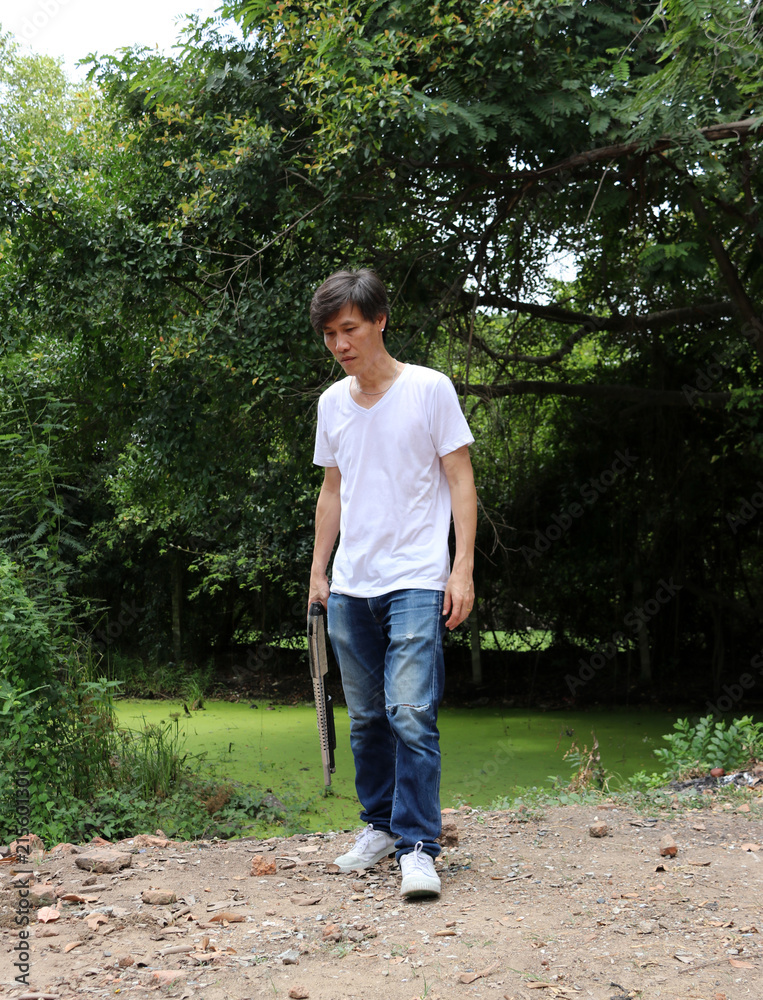 The gunner man holding long gun in hand with white shirt and denim jean on green tree background. The gunner man holding long gun in hand with white shirt and denim jean on green tree background.