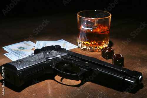Glass of whiskey or cognac, gun, playing cards with money on the black mirror table. The concept of criminal.