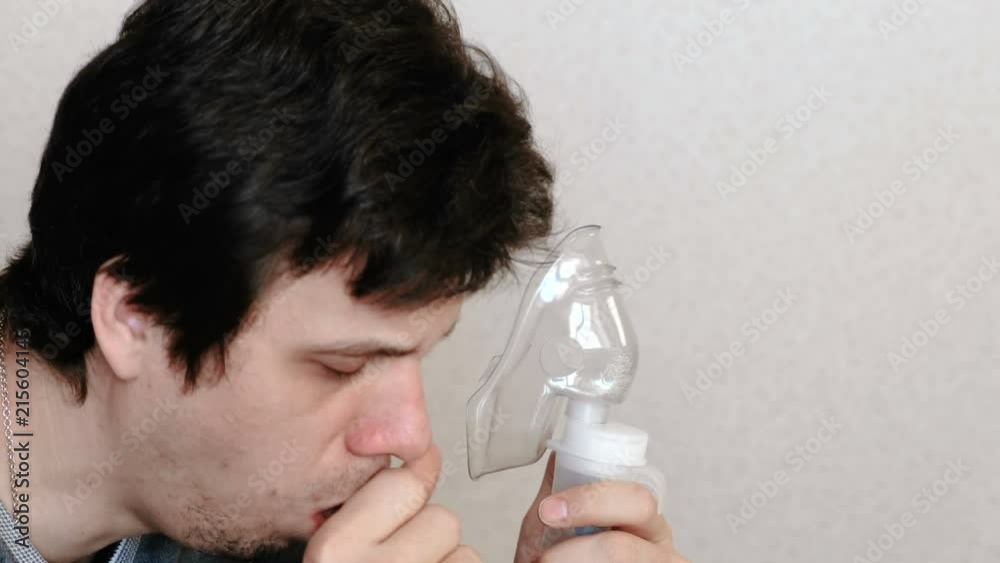 Use nebulizer and inhaler for the treatment. Young man inhaling through inhaler mask and coughs. Side view. Use nebulizer and inhaler for the treatment. Young man inhaling through inhaler mask and coughs. Side view.