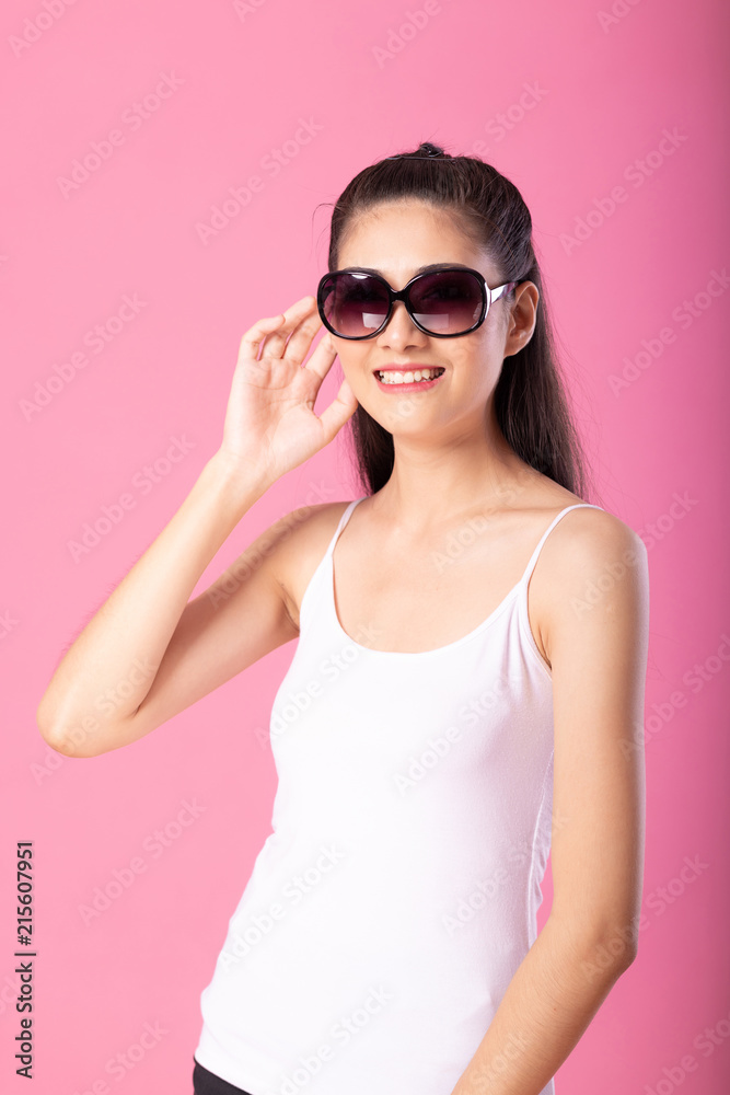 Portrait of a smiling attractive woman in white tanktop outfit and sunglasses posing while standing and smiling at camera isolated over pink background. Portrait of a smiling attractive woman in white tanktop outfit and sunglasses posing while standing and smiling at camera isolated over pink background.