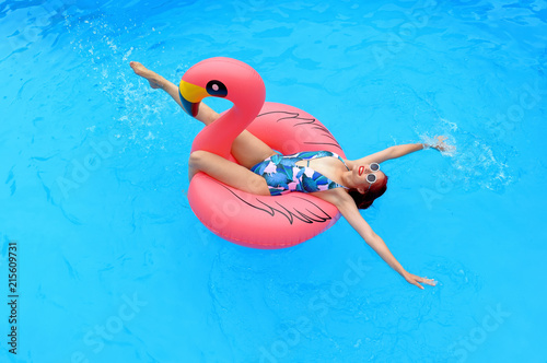 A young girl wearing a bathing suit and a white summer hat sits relaxed in an inflatable swim ring. She poses in  a pool smiling at the camera.