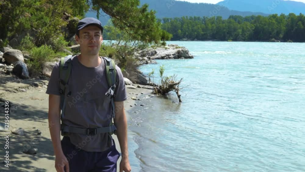 A young tourist with a backpack waving his hand to his friends. In the background a river, forest, mountains, blue sky. A young tourist with a backpack waving his hand to his friends. In the background a river, forest, mountains, blue sky.