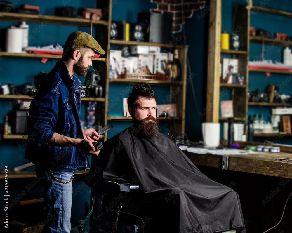 Barber styling hair of bearded client with comb and clipper. Hipster client getting haircut. Haircut concept. Barber with hair clipper works on hairstyle for man with beard, barbershop background