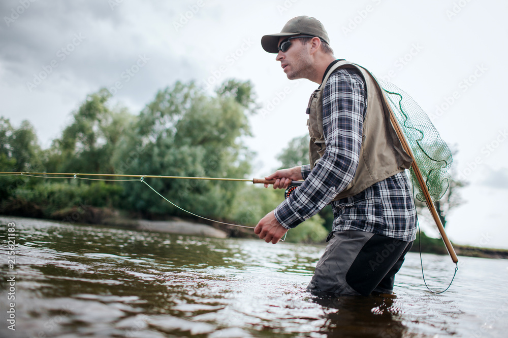 Fisherman in action. He stands in water and holds fly rod in one hand and spoon in the other one. Also adult has a fishing net on the back. Green trees are at the edge of water. Fisherman in action. He stands in water and holds fly rod in one hand and spoon in the other one. Also adult has a fishing net on the back. Green trees are at the edge of water.
