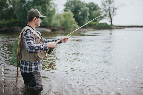 Serious man is standing in water and fishing. He has spinning in hands and fishing net on the back. Guy wears vest, waders, shirt and cap.