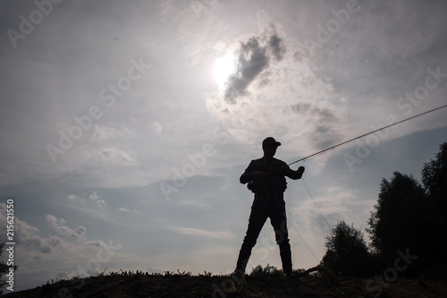 Dark picture of fisherman standsat the edge of water. He holds fly rod in hads. He is fishing. Sun is behind cloud.