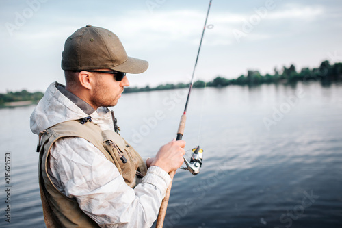 A picture of man in cap, sunglasses and vest standing in shallow and looking at fly rod. He is using non-inertial reel. Guy is concentrated.