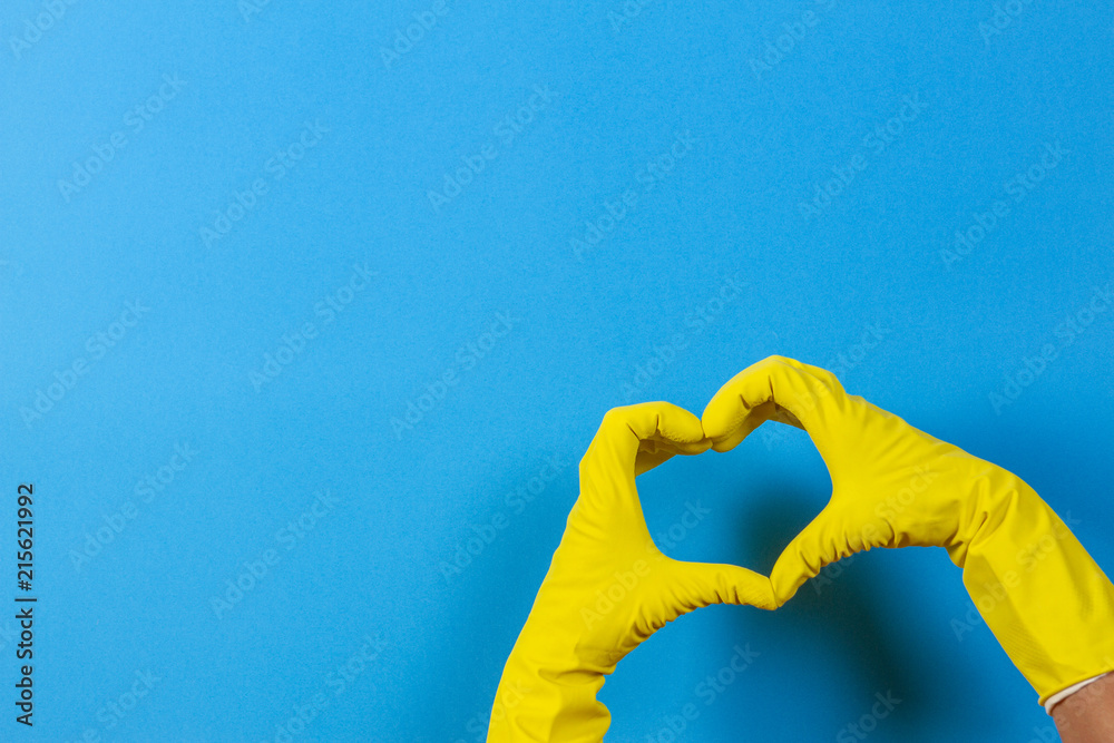 Hands in yellow rubber gloves making heart shape with fingers, on blue background - obrazy, fototapety, plakaty 
