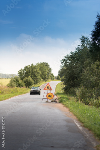 A warning sign on the road runs ahead. Road sign. Road repair and small speed 40. kilometers per hour. A big hole in the asphalt rural road. trees and field under the blue sky