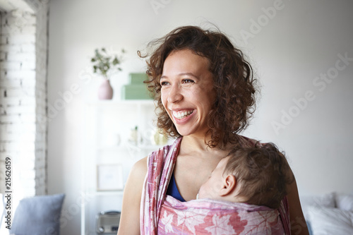 Horizontal shot of emotional young mixed race woman with wavy hair doing her daily routine at home while her little son is having nap in one shouldered baby carrier, posing in light room interior photo