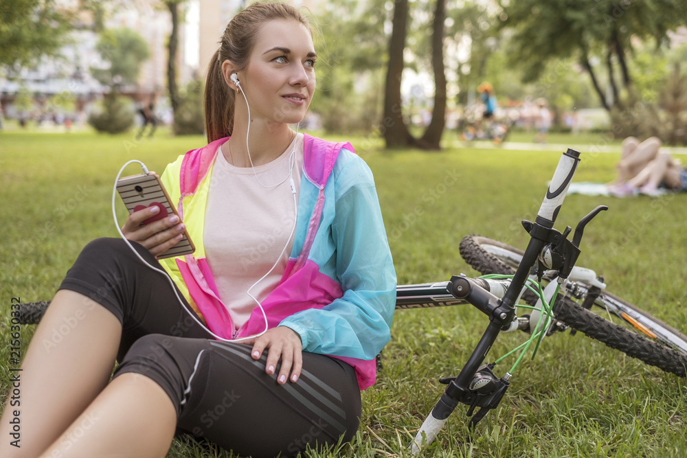 Fototapeta premium Young woman in sports wear relaxing in park after riding a bicycle. Fit, slim brunette woman with cheerful smile. Summer weather, outdoors leisure.