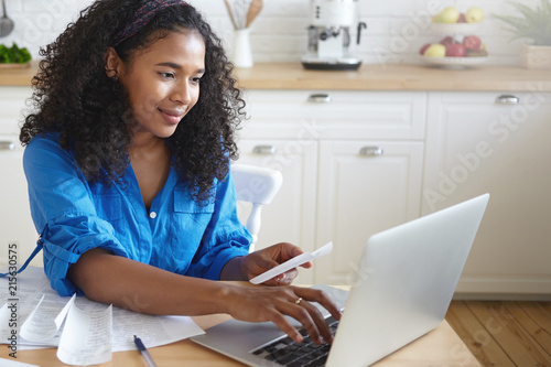 Candid shot of focused beautiful young African housewife wearing headband and casual shirt paying utility bills online using laptop computer, sitting at kitchen table with papers and smiling photo