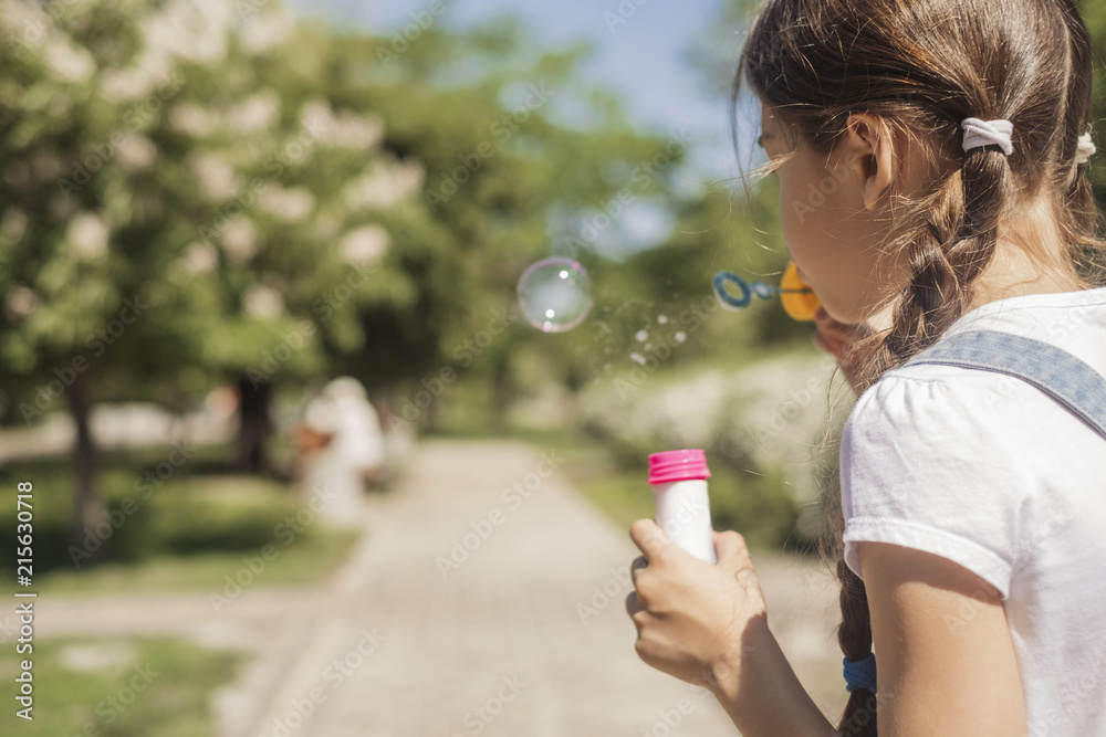 Cute little 10 years old girl in casual outfit playing at park in warm summer day. She blows soap bubbles. Copy space