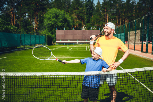 Positive male athlete with cheerful boy playing tennis. Glad kid pointing racket while situating on court near him