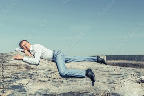 A young man in a white shirt and blue trousers is lying on a log, sleeping in the fresh air against the blue sky.