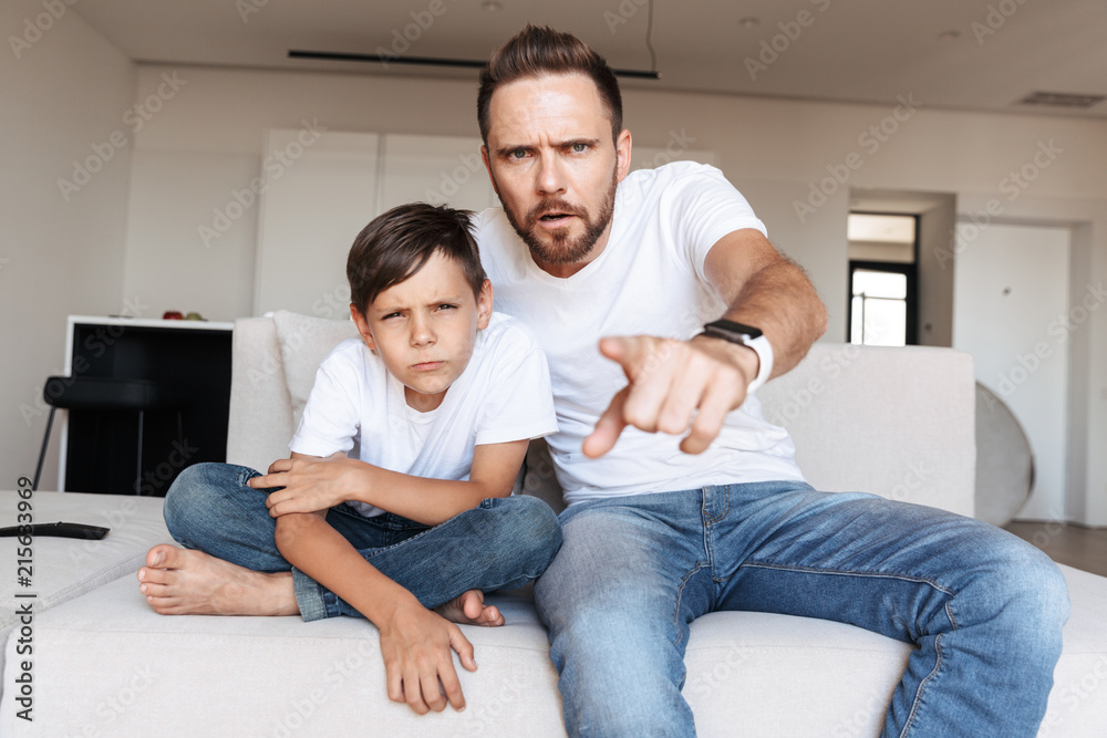 Portrait of serious concentrated man 30s and boy 8-10 pointing finger at you, while sitting on sofa in apartment Portrait of serious concentrated man 30s and boy 8-10 pointing finger at you, while sitting on sofa in apartment