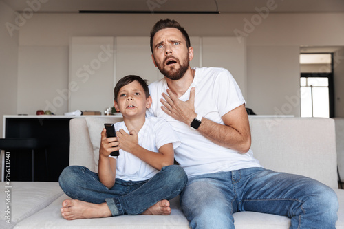 Image of outraged emotional man 30s with son 8-10 watching tv, while sitting on sofa in room
