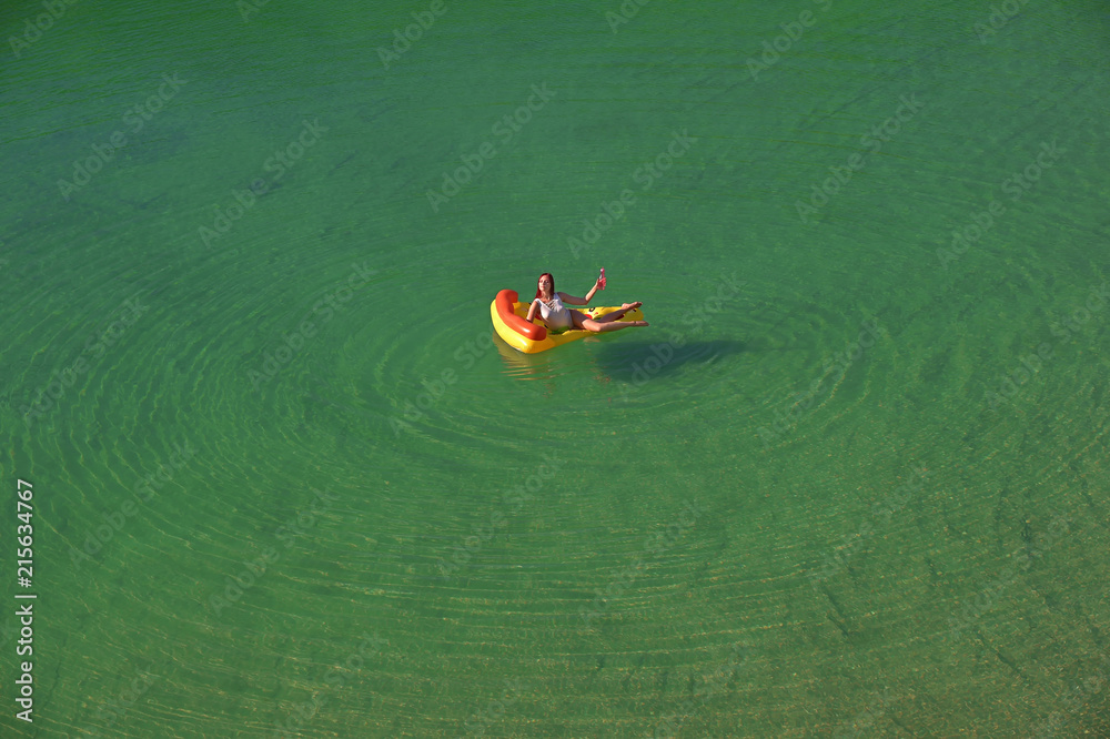 Fototapeta premium A young woman spends leisure time on an inflatable sun bed in the middle of a green lake. She wears a white swim suit and enjoys the summer sun as she relaxes on the sun bed. 
