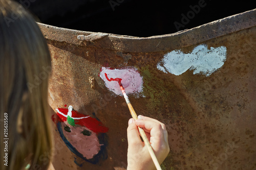 A child's hand holds a brush. Point of view. The child paints a color paint on an iron barrel.
