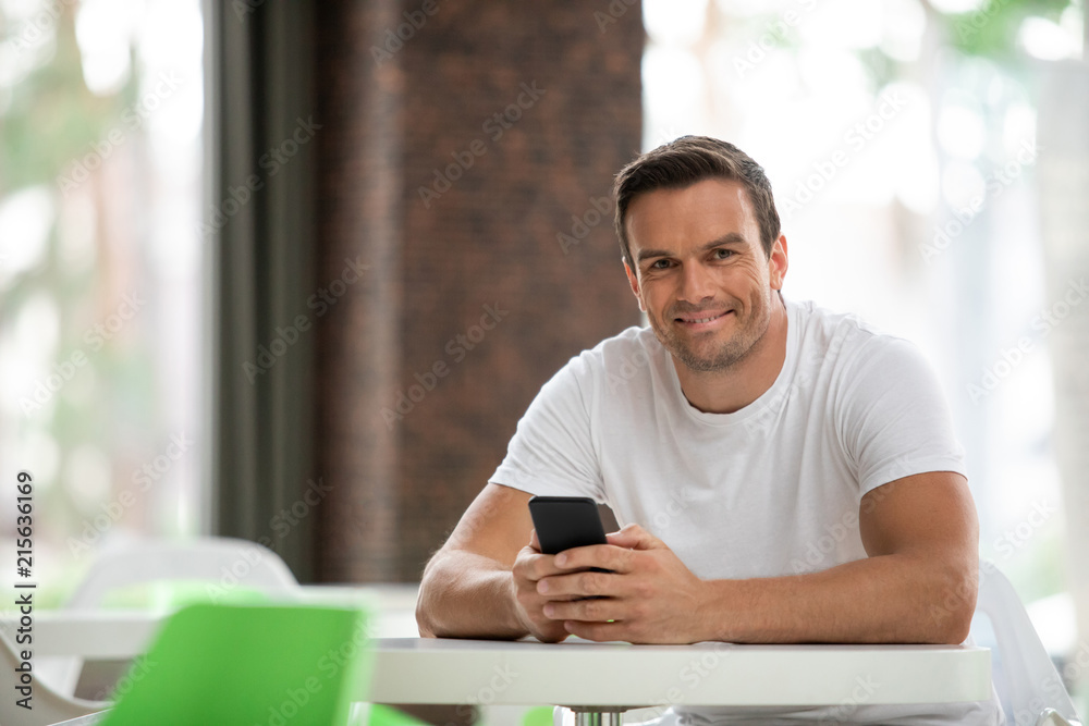 Waist up portrait of delighted male sitting at table and surfing net with joy. He is holding smartphone while waiting for friend with smile. Copy space in left side Waist up portrait of delighted male sitting at table and surfing net with joy. He is holding smartphone while waiting for friend with smile. Copy space in left side