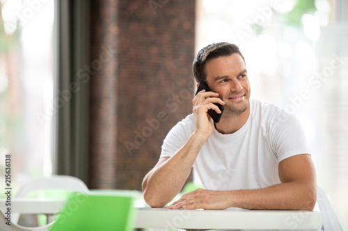 Waist up portrait of smiling man sitting at table with mobile in hands. He is talking to his friend while waiting for order with content. Copy space in left side