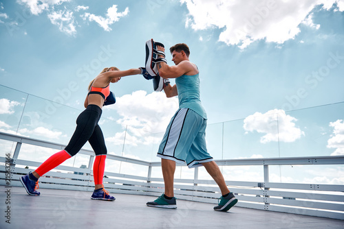 Fototapeta Naklejka Na Ścianę i Meble -  Instructor is having workout with slim lady on terrace. He is standing and using safety pads while she is kicking with gloves. Exercising for defending yourself concept