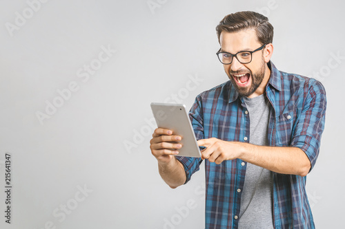 OMG! Great news! Surprised young man in plaid shirt standing and using tablet over grey background.