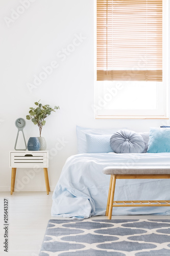 Window with wooden blinds in white bedroom interior with bed with light blue bedclothes, carpet with pattern and bedside table with clock and plant